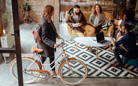 A newly hired temporary creative worker wheels her bike into the office and is greeted by her new coworkers